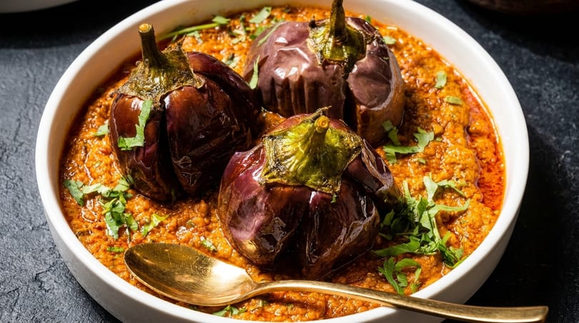 Three whole eggplants in a rich, orange-red curry sauce, garnished with fresh cilantro and a golden spoon.