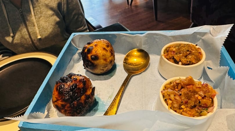 Two charred bread rolls, a golden spoon, and two bowls of chutney on a blue tray.